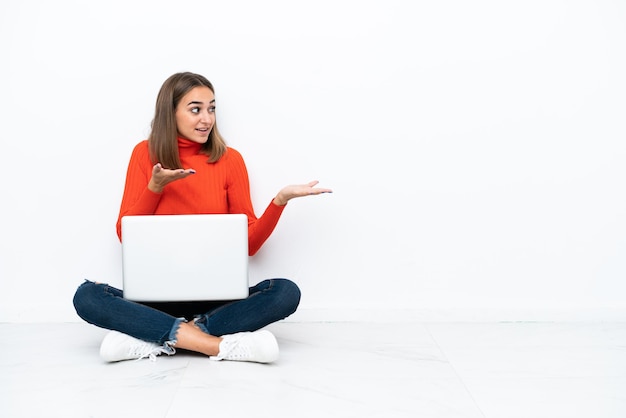 Giovane donna caucasica seduta sul pavimento con un laptop con espressione facciale a sorpresa