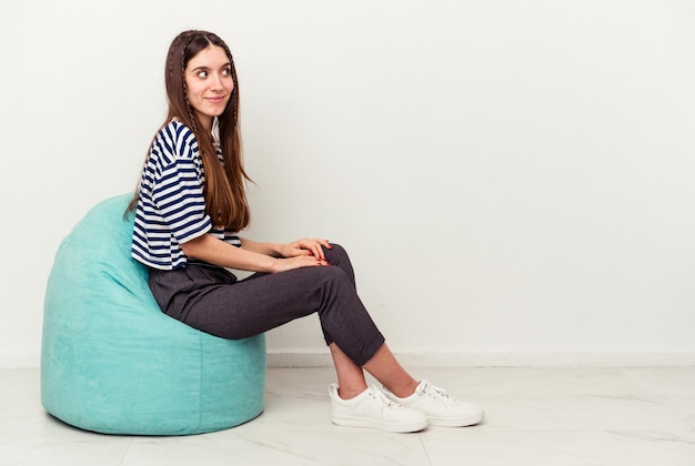 Young caucasian woman sitting on a puff isolated on white background looks aside smiling, cheerful and pleasant.