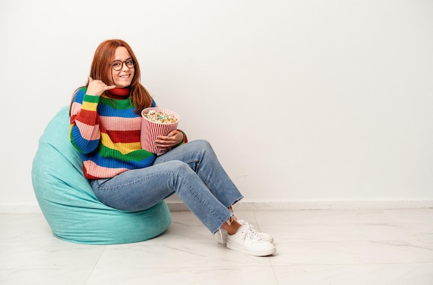 Photo young caucasian woman sitting on puff with popcorns isolated on white background showing a mobile phone call gesture with fingers.