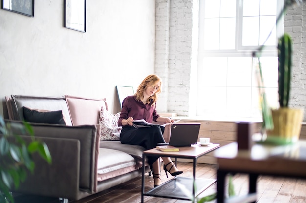 Giovane donna caucasica seduta sul divano, lavorando presso un ufficio moderno. sul tavolo ci sono laptop, notebook e tazza di caffè. concetto di affari