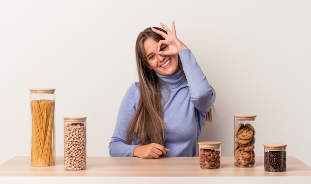 Young caucasian woman sitting at a table with food pot isolated on white background excited keeping ok gesture on eye.