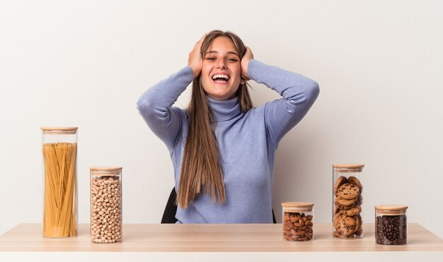 Young caucasian woman sitting at a table with food pot isolated on white background laughs joyfully keeping hands on head. Happiness concept.