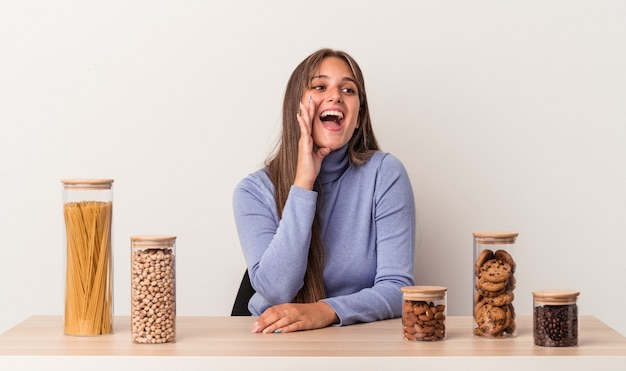 Young caucasian woman sitting at a table with food pot isolated on white background shouting and holding palm near opened mouth.