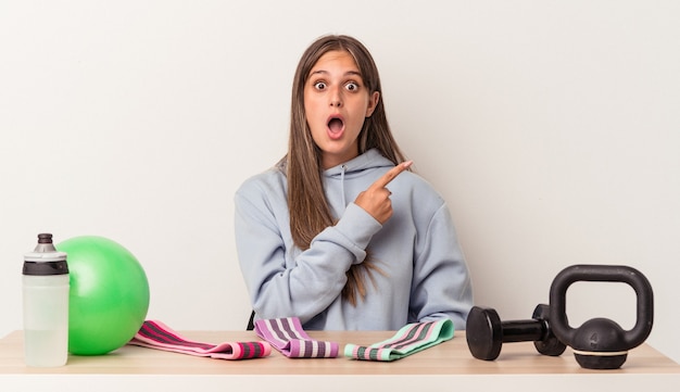 Young caucasian woman sitting at a table with sport equipment isolated on white background pointing to the side