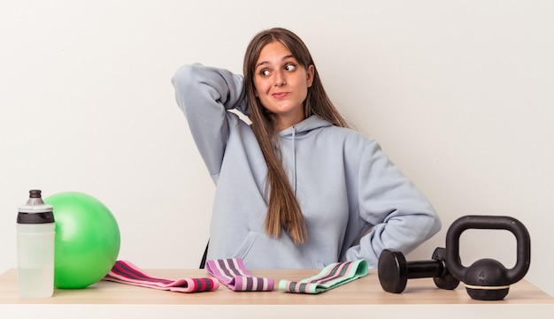 Young caucasian woman sitting at a table with sport equipment isolated on white background touching back of head, thinking and making a choice.