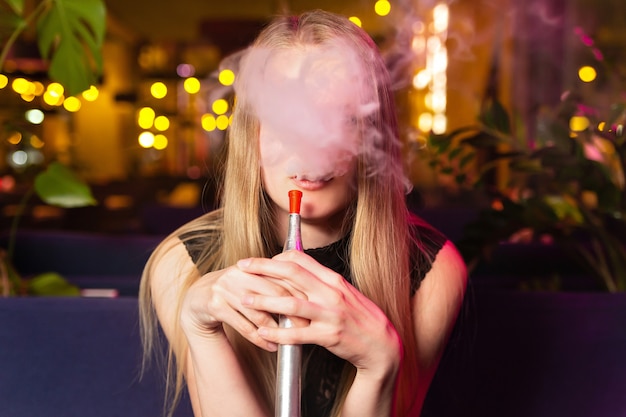 Young caucasian woman smokes a hookah or shisha in the night club or bar smoke. Unrecognizable face behind smoke