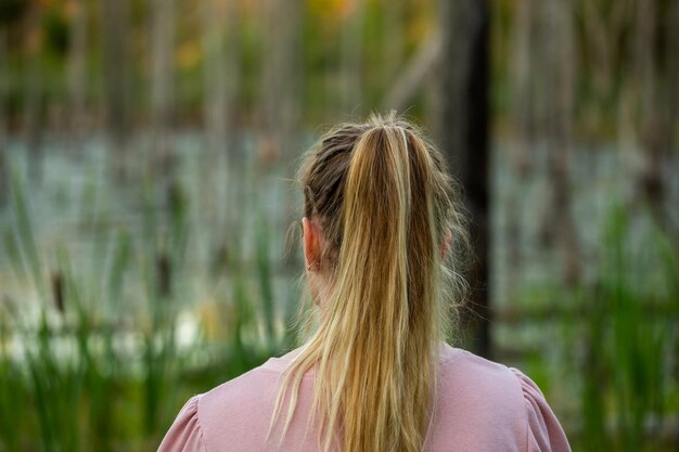 背景にぼやけた乾燥した木とキャタイと夕方の沼でカメラに戻って立っている若い白人女性