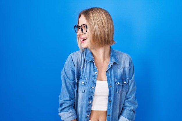 Young caucasian woman standing over blue background looking away to side with smile on face, natural expression. laughing confident.