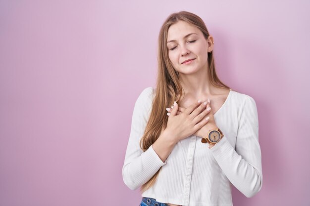 Young caucasian woman standing over pink background smiling with hands on chest with closed eyes and grateful gesture on face health concept