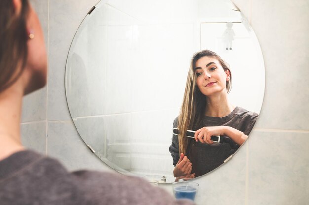 Young caucasian woman straightening her hair with a hair straightenering on the mirror at the bathroom.