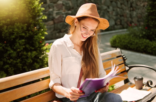 Young Caucasian woman student in casual clothing reading esse in folder while sitting on bench at park.