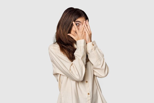 Young Caucasian woman in studio setting blink through fingers frightened and nervous