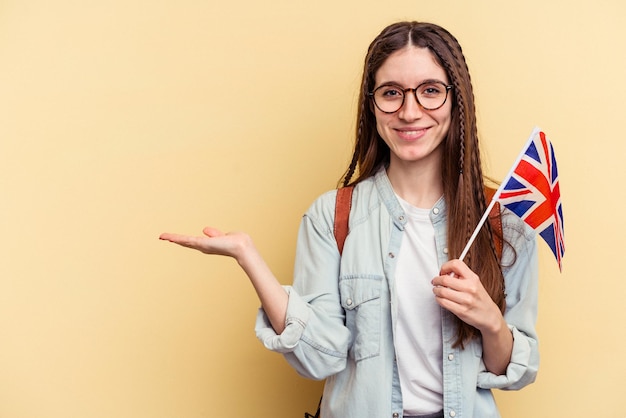 Young caucasian woman studying English isolated on yellow background showing a copy space on a palm and holding another hand on waist.