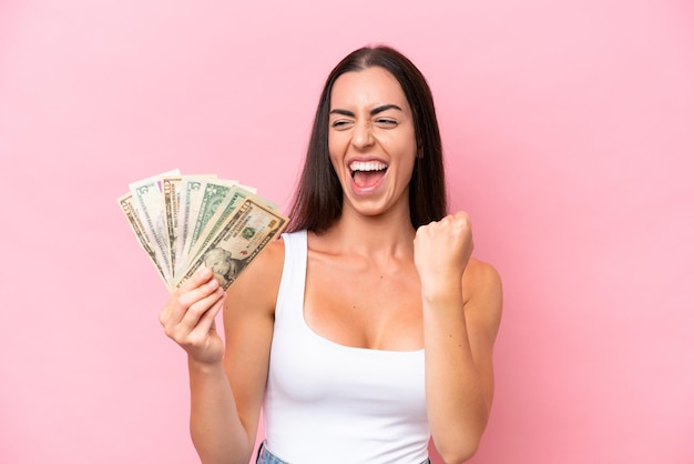 Young caucasian woman taking a lot of money isolated on pink background celebrating a victory