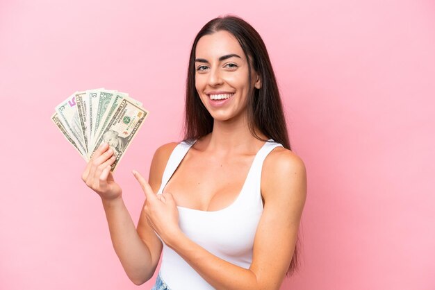 Young caucasian woman taking a lot of money isolated on pink background and pointing it