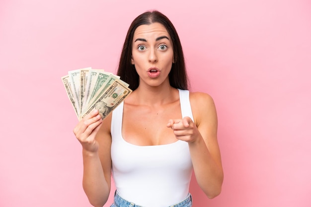 Young caucasian woman taking a lot of money isolated on pink background surprised and pointing front