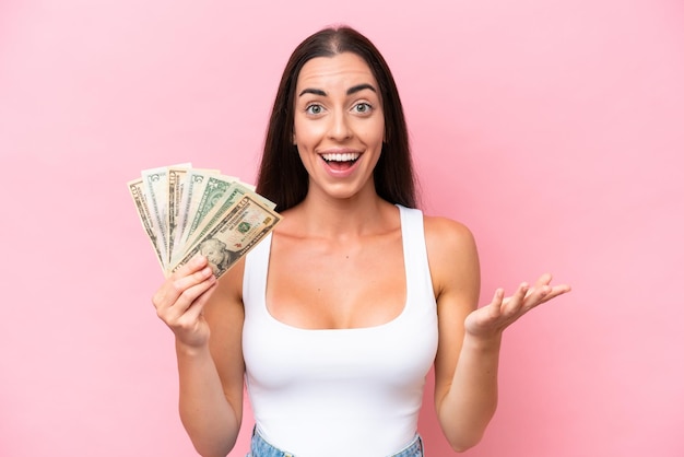 Young caucasian woman taking a lot of money isolated on pink background with shocked facial expression