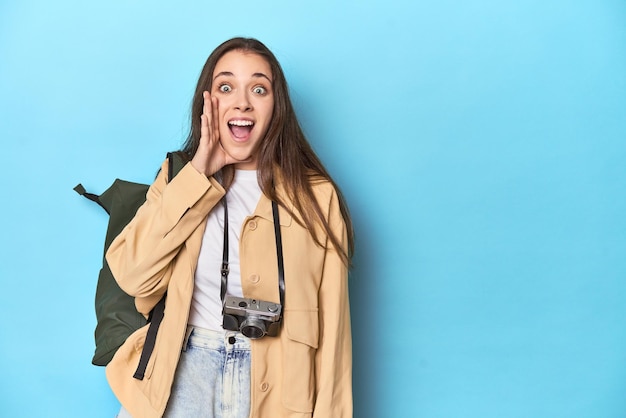 Young caucasian woman traveler with camera and backpack shouting and holding palm near opened mouth