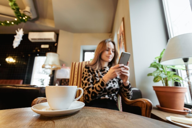 携帯電話を使用して、カフェのテーブルに座って、前景にコーヒーを飲む若い白人女性