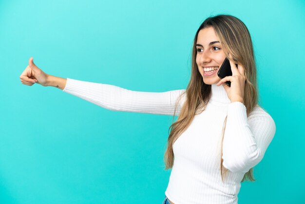 Young caucasian woman using mobile phone isolated on blue background giving a thumbs up gesture