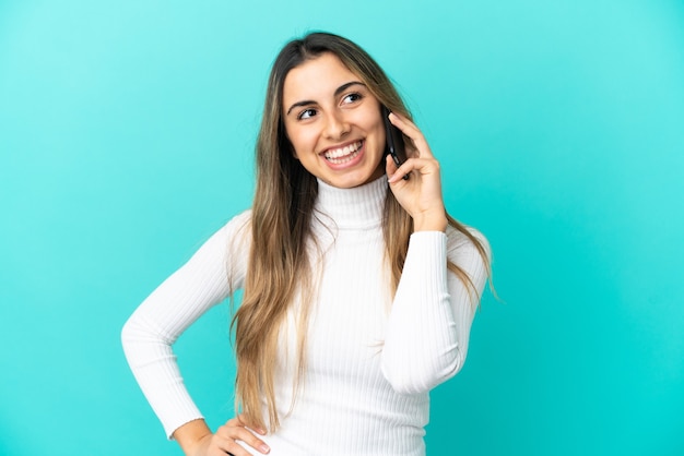 Foto giovane donna caucasica utilizzando il telefono cellulare isolato su sfondo blu in posa con le braccia all'anca e sorridente