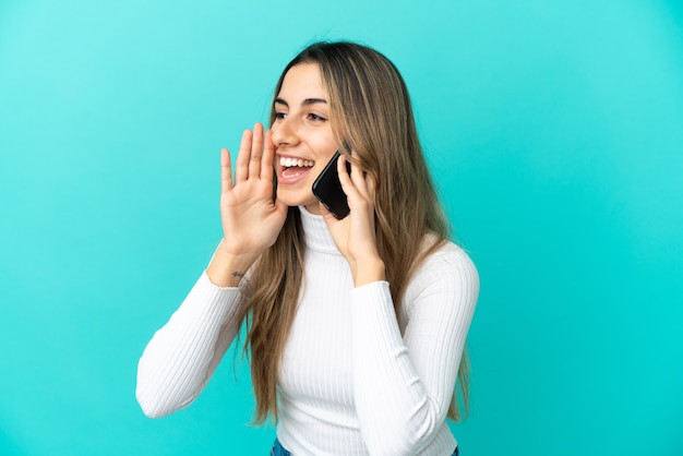 Young caucasian woman using mobile phone isolated on blue background shouting with mouth wide open to the side