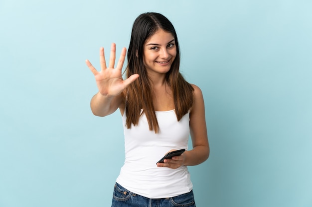 Giovane donna caucasica utilizzando il telefono cellulare isolato su blu contando cinque con le dita