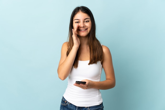 Giovane donna caucasica utilizzando il telefono cellulare isolato sulla parete blu che grida con la bocca spalancata