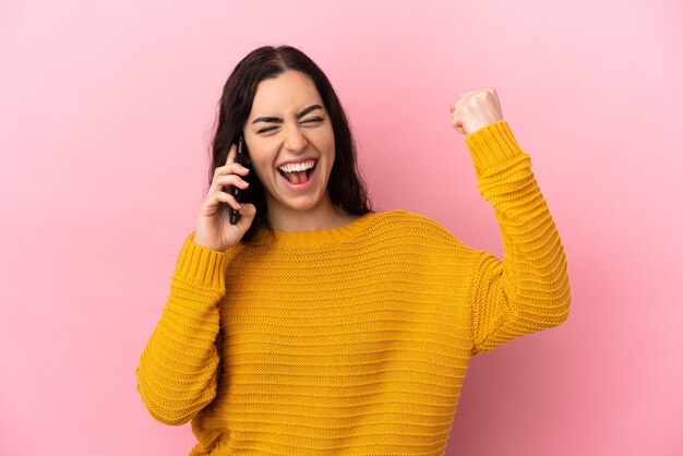 勝利を祝うピンクの背景に分離された携帯電話を使用して若い白人女性