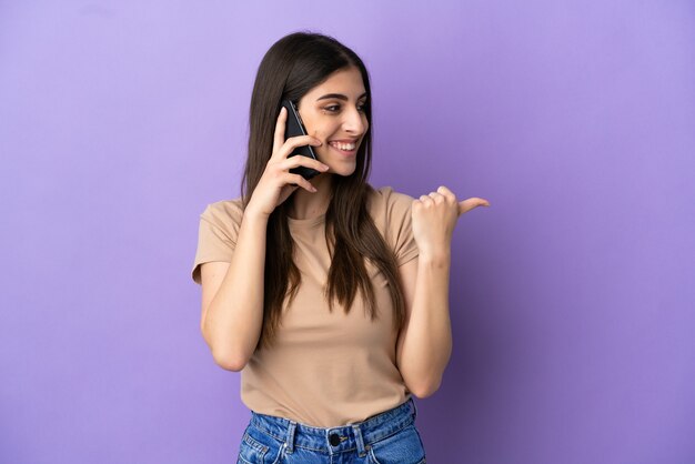 제품을 제시하기 위해 측면을 가리키는 보라색 배경에 격리된 휴대 전화를 사용하는 젊은 백인 여성