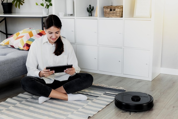 Photo young caucasian woman using tablet chatting playing while robot vacuum cleaner is cleaning floor