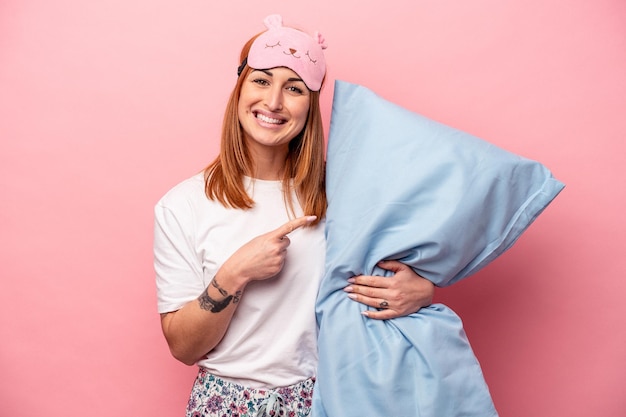 写真 ピンクの背景に分離された枕を保持しているパジャマを着ている若い白人女性は、笑顔で脇を指して、空白のスペースで何かを示しています。