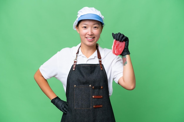 Young caucasian woman wearing an apron and serving fresh cut meat over isolated background posing with arms at hip and smiling