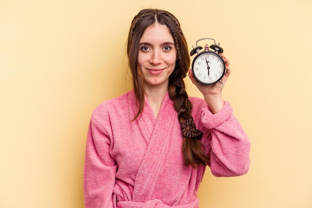 Young caucasian woman wearing a bathrobe holding a alarm clock isolated on yellow background happy smiling and cheerful