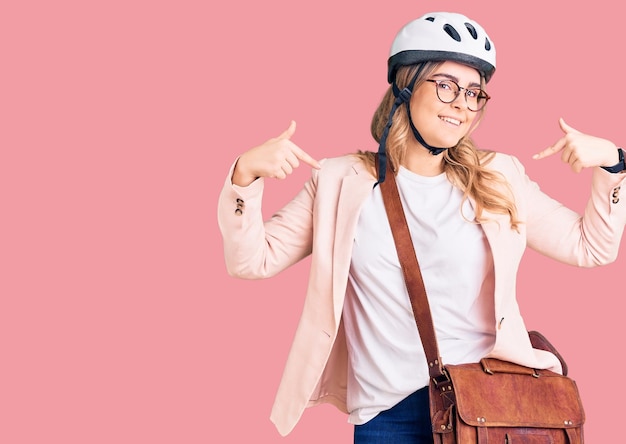 Young caucasian woman wearing bike helmet and leather bag looking confident with smile on face, pointing oneself with fingers proud and happy.