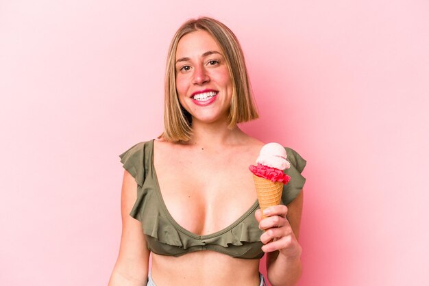 Young caucasian woman wearing a bikini and holding an ice cream isolated on pink background happy smiling and cheerful