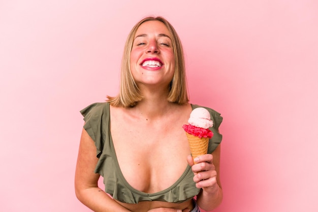 Young caucasian woman wearing a bikini and holding an ice cream isolated on pink background laughing and having fun