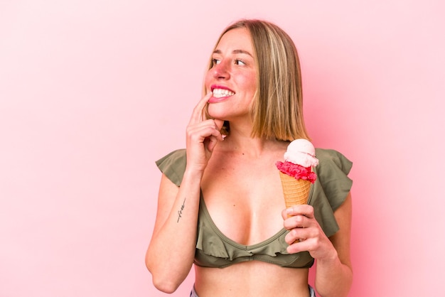 Young caucasian woman wearing a bikini and holding an ice cream isolated on pink background relaxed thinking about something looking at a copy space