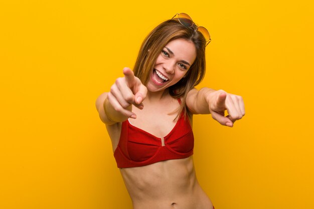 Young caucasian woman wearing bikini and sunglasses cheerful smiles pointing to front.