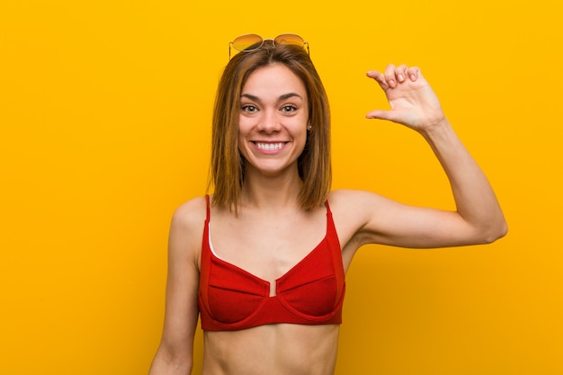 Bikini d'uso ed occhiali da sole della giovane donna caucasica che tengono qualcosa poco con gli indici, sorridenti e sicuri.