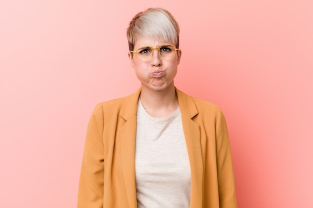 Young caucasian woman wearing a casual business clothes blows cheeks, has tired expression