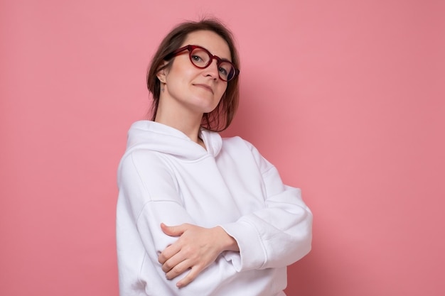 Giovane donna caucasica che indossa una felpa casual con felpa con cappuccio con le braccia incrociate guardando la telecamera. studio girato sul muro rosa.