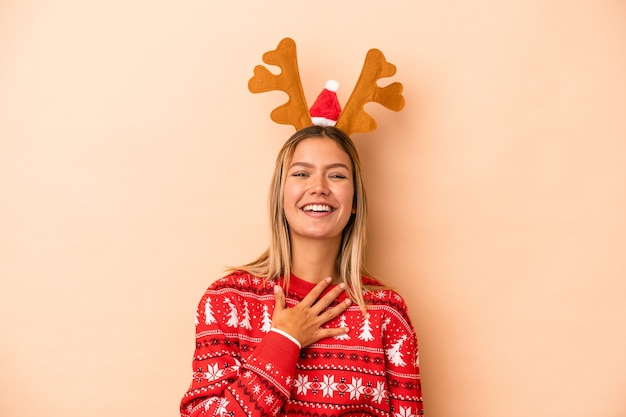 Young caucasian woman wearing a christmas reindeer hat isolated on beige background laughs out loudly keeping hand on chest.