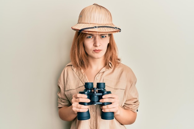 写真 探検家の帽子をかぶった若い白人女性は、双眼鏡で懐疑的で神経質になり、問題のために眉をひそめています。ネガティブな人。