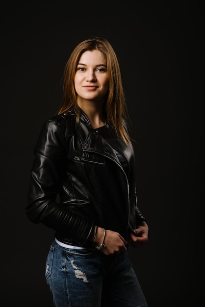 Молодая кавказская женщина в кожаной куртке и джинсах позирует на черном фоне