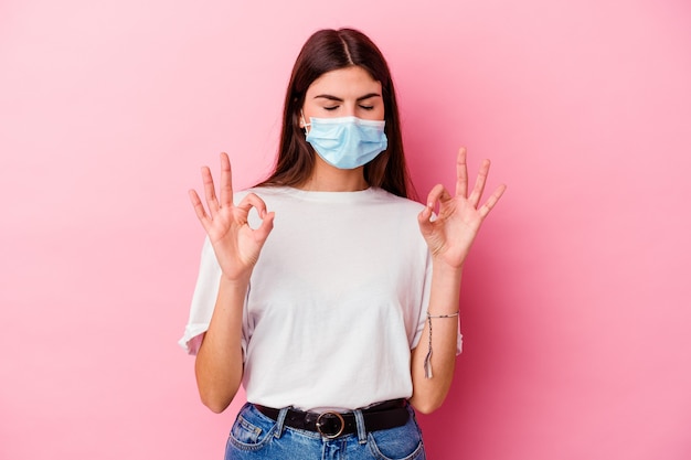 Young caucasian woman wearing a mask for virus isolated on pink background relaxes after hard working day, she is performing yoga.