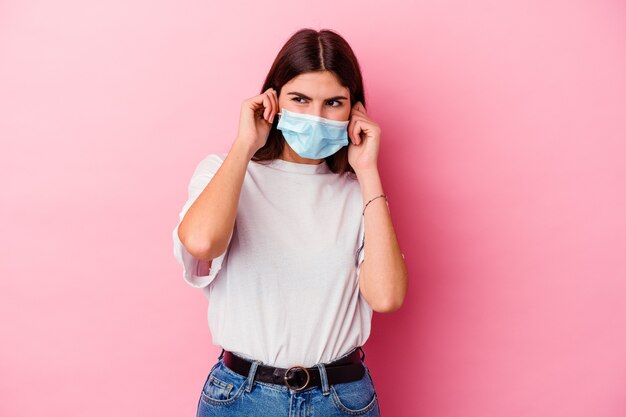 Young caucasian woman wearing a mask for virus isolated on pink wall covering ears with hands