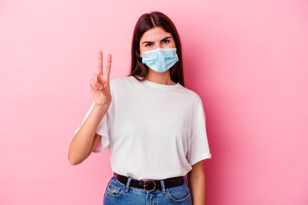 Young caucasian woman wearing a mask for virus on pink showing victory sign and smiling broadly.