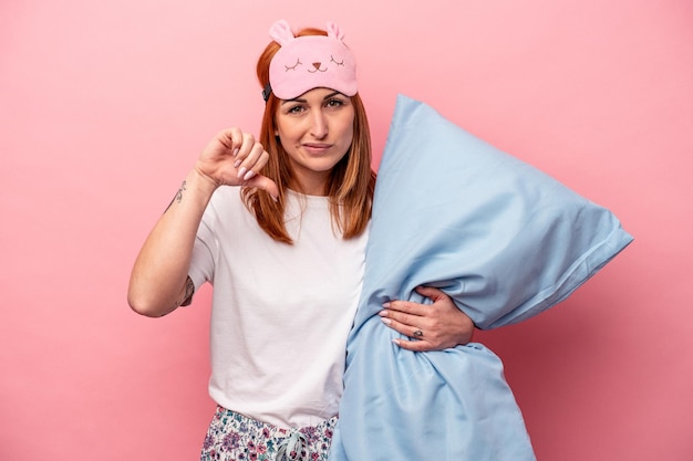 Young caucasian woman wearing a pajama holding pillow isolated on pink background showing a dislike gesture thumbs down Disagreement concept