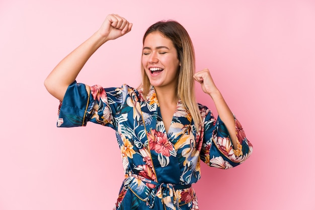 Young caucasian woman wearing pajamas celebrating a special day, jumps and raise arms with energy.